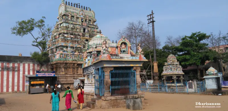 A colourful temple tower rising behind a small shrine in a sandy courtyard, with visitors walking past