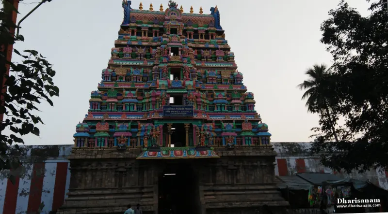 A tall tiered gopuram with a name board, a red and white striped wall beside it, and trees around